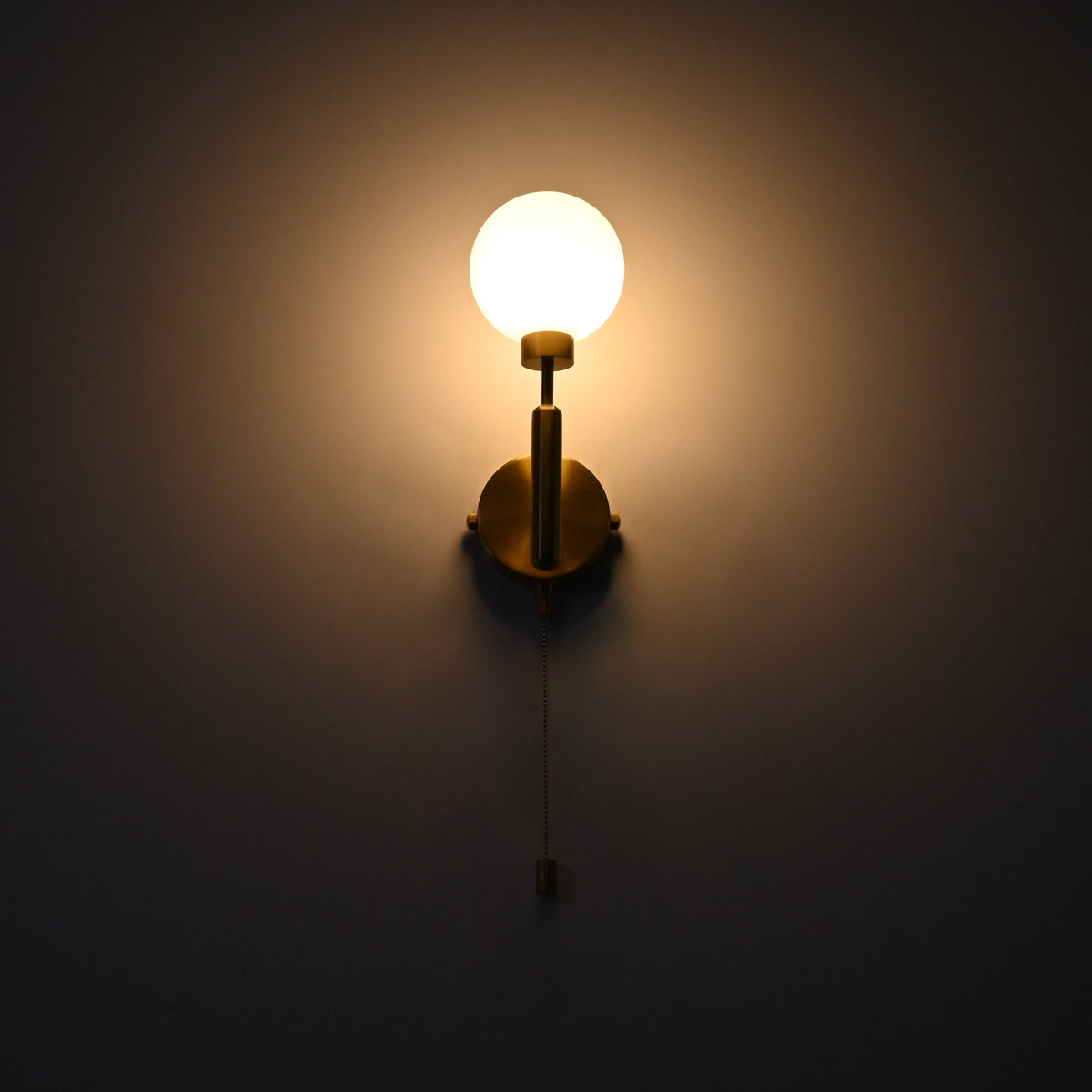 Raw brass wall sconce with a round frosted glass globe illuminated warmly, mounted on a dark wall with a hanging pull chain.