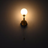 Raw brass wall sconce with a round frosted glass globe illuminated warmly, mounted on a dark wall with a hanging pull chain.