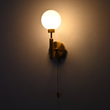 Raw brass wall sconce with a round frosted glass globe shade, illuminated, featuring a pull chain switch against a dark wall.