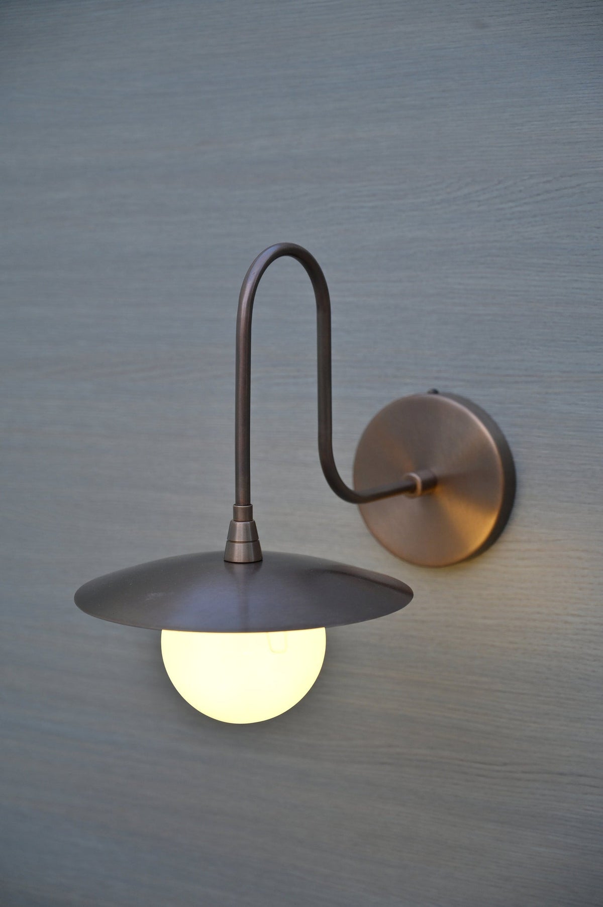 Side view of industrial brass wall sconce with matte finish, curved arm, and glowing glass globe shade.