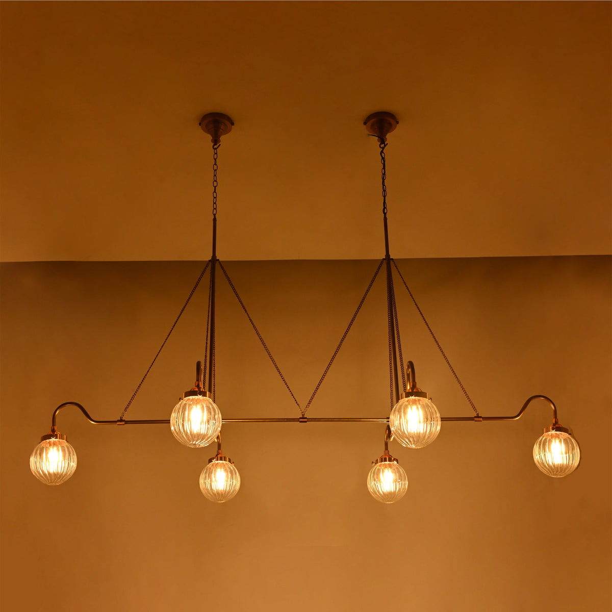 Linear 6-light ribbed glass globe chandelier shown in a warm bedroom interior with brass frame and ambient ceiling lighting