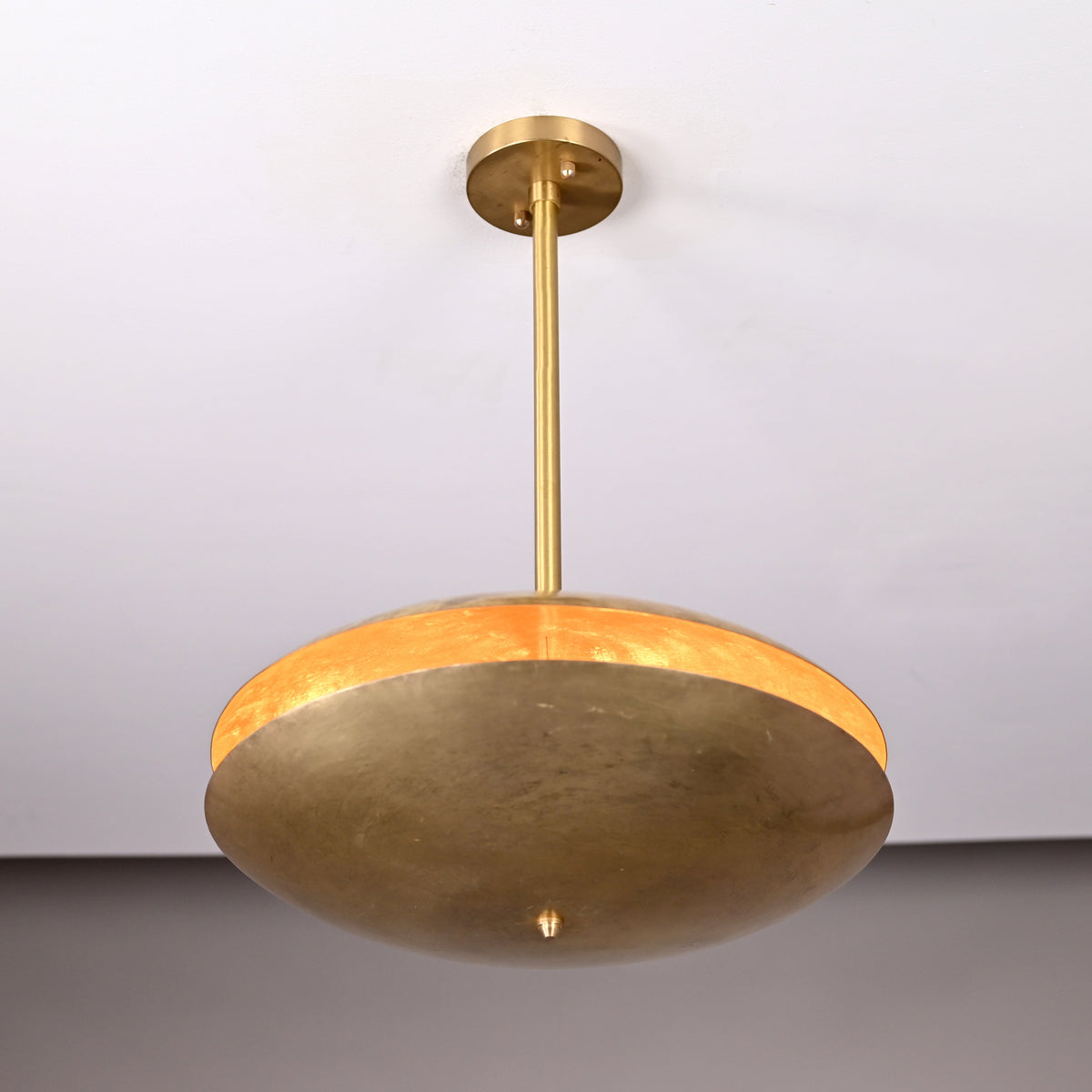 Modern brass disc pendant light viewed from a low upward angle, displaying warm inner glow and handcrafted hammered surface.