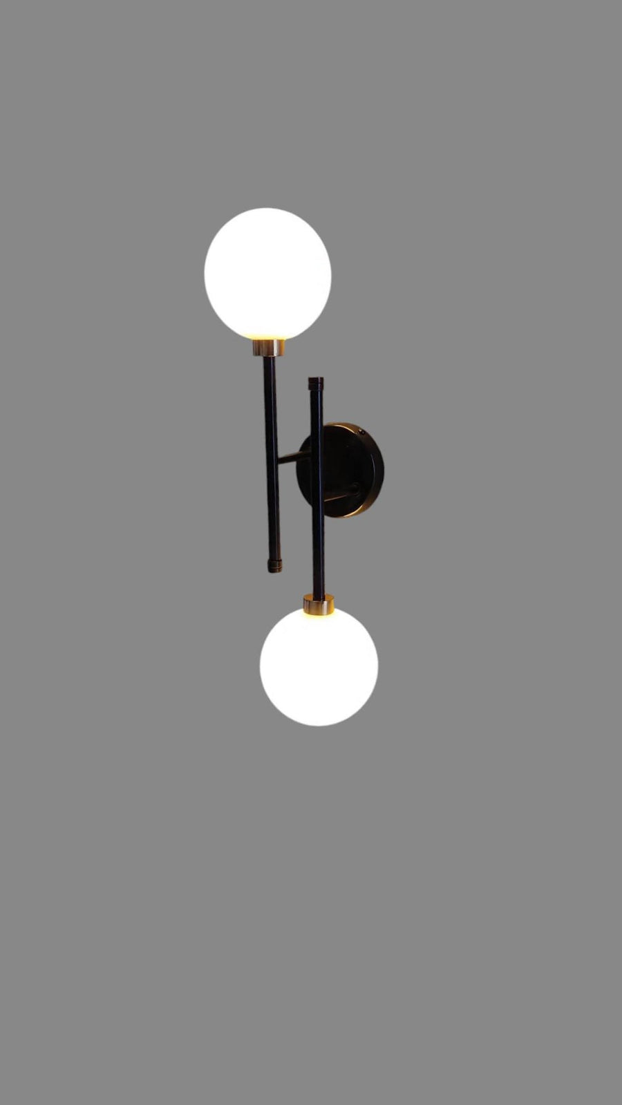 Illuminated double globe wall light in black and brass finish, mounted on wall providing warm ambient glow above indoor plants.