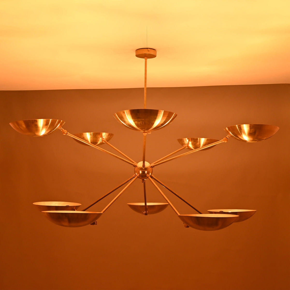 Sculptural radial brass chandelier with ten bowl-shaped uplights glowing warmly against a dark background.