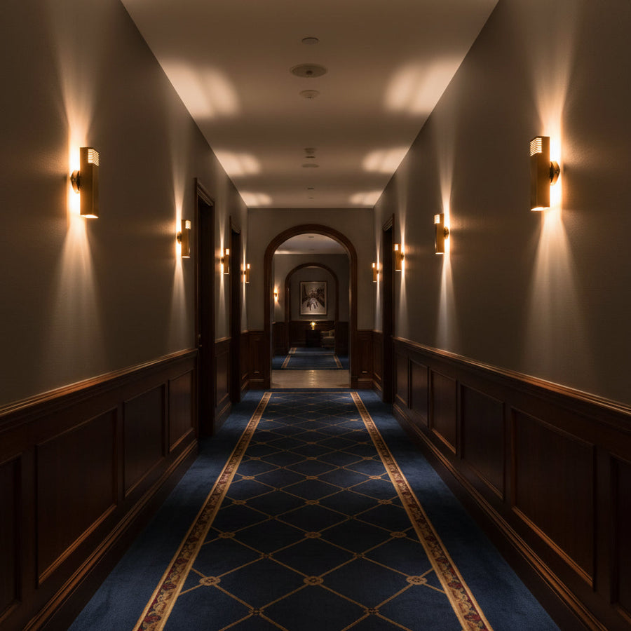 Modern brass tube wall sconce shown illuminated with decorative slotted detail and warm light pattern on the wall