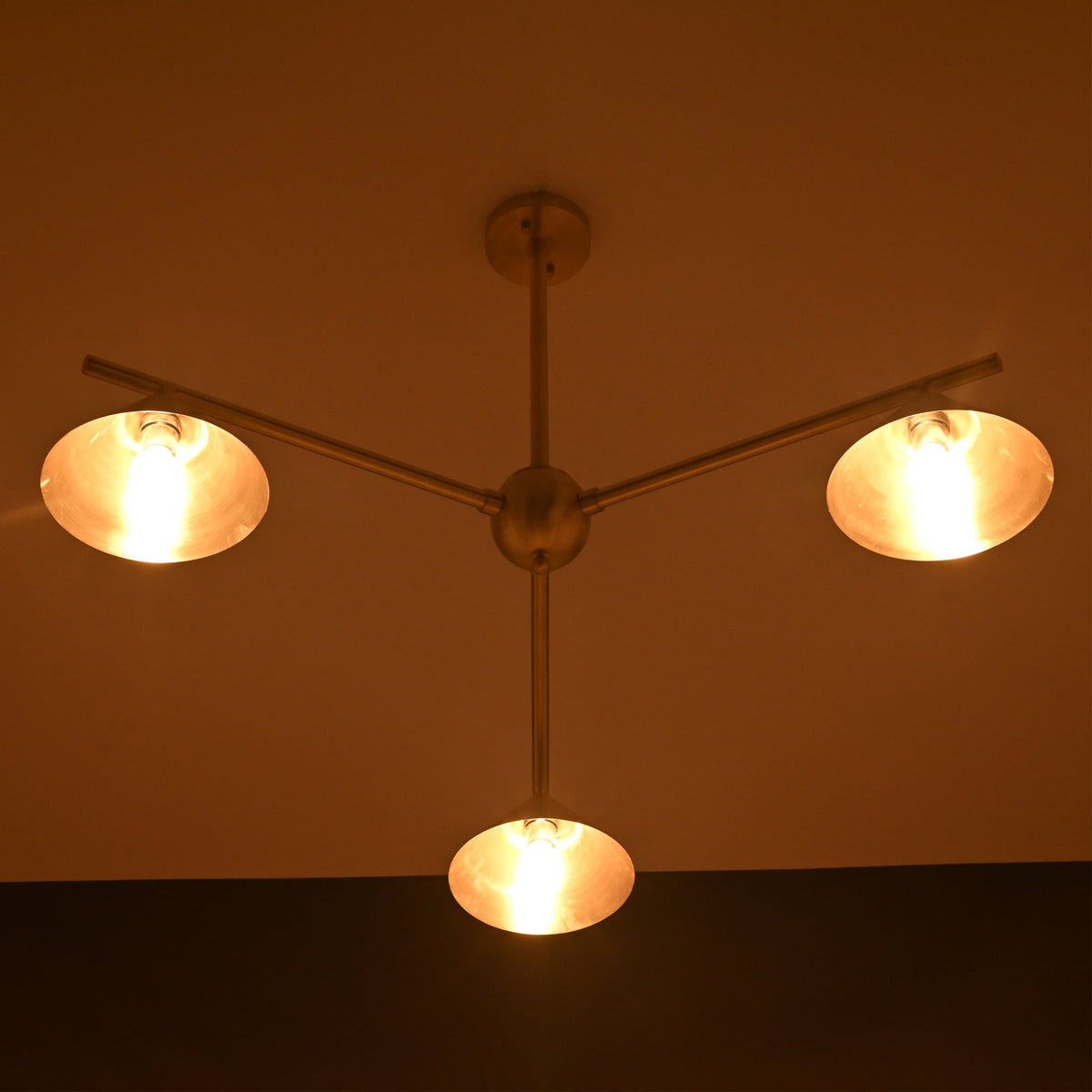 Brushed brass 3-light chandelier illuminated in warm ambient lighting, showcasing cone shades in a dark room.